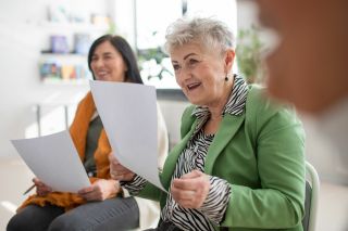Vrouw met grijs haar en een groen jasje houdt een papier vast en is aan het oefenen met zingen